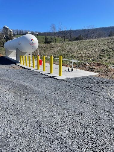 Large white propane tank on concrete pad, yellow bollards, gravel driveway, and blue sky.