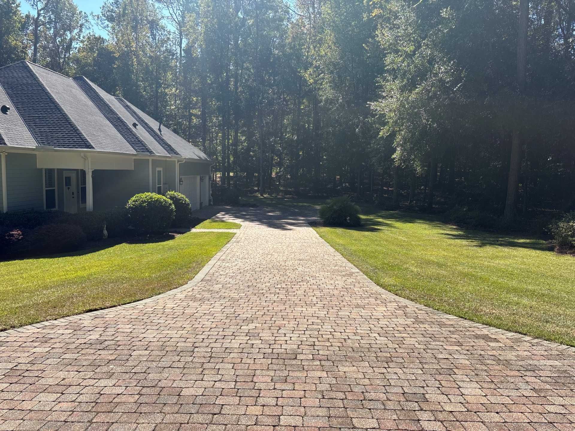 A brick driveway leading to a light-colored house surrounded by trees on a sunny day.