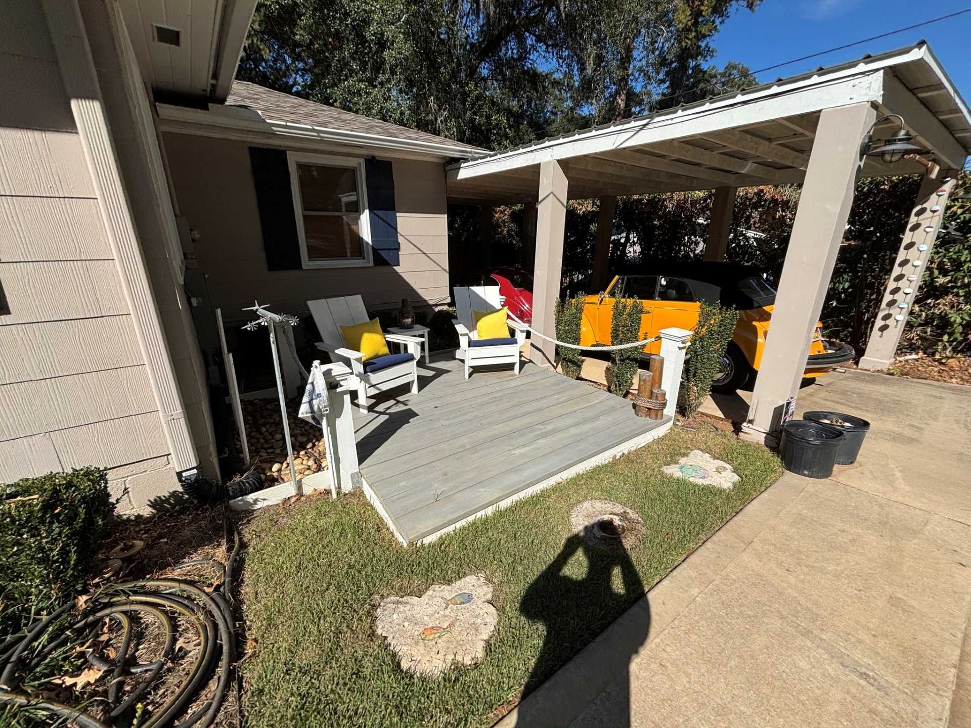 A small wooden patio with two chairs and yellow cushions sits next to a house with a carport and a parked yellow car.