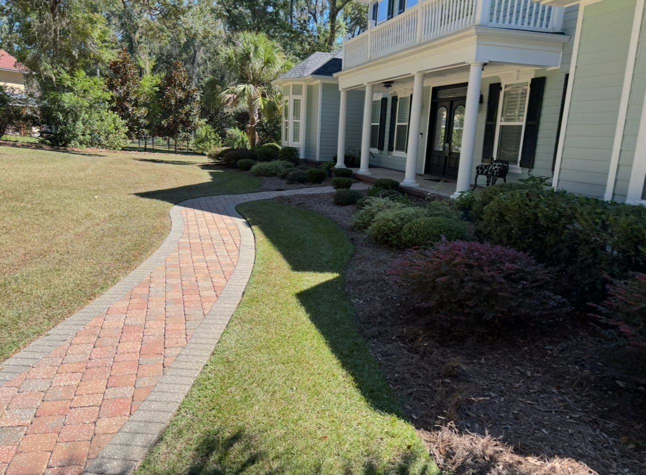 A curved red brick pathway leading to the front porch of a light green house with white columns and a front lawn.