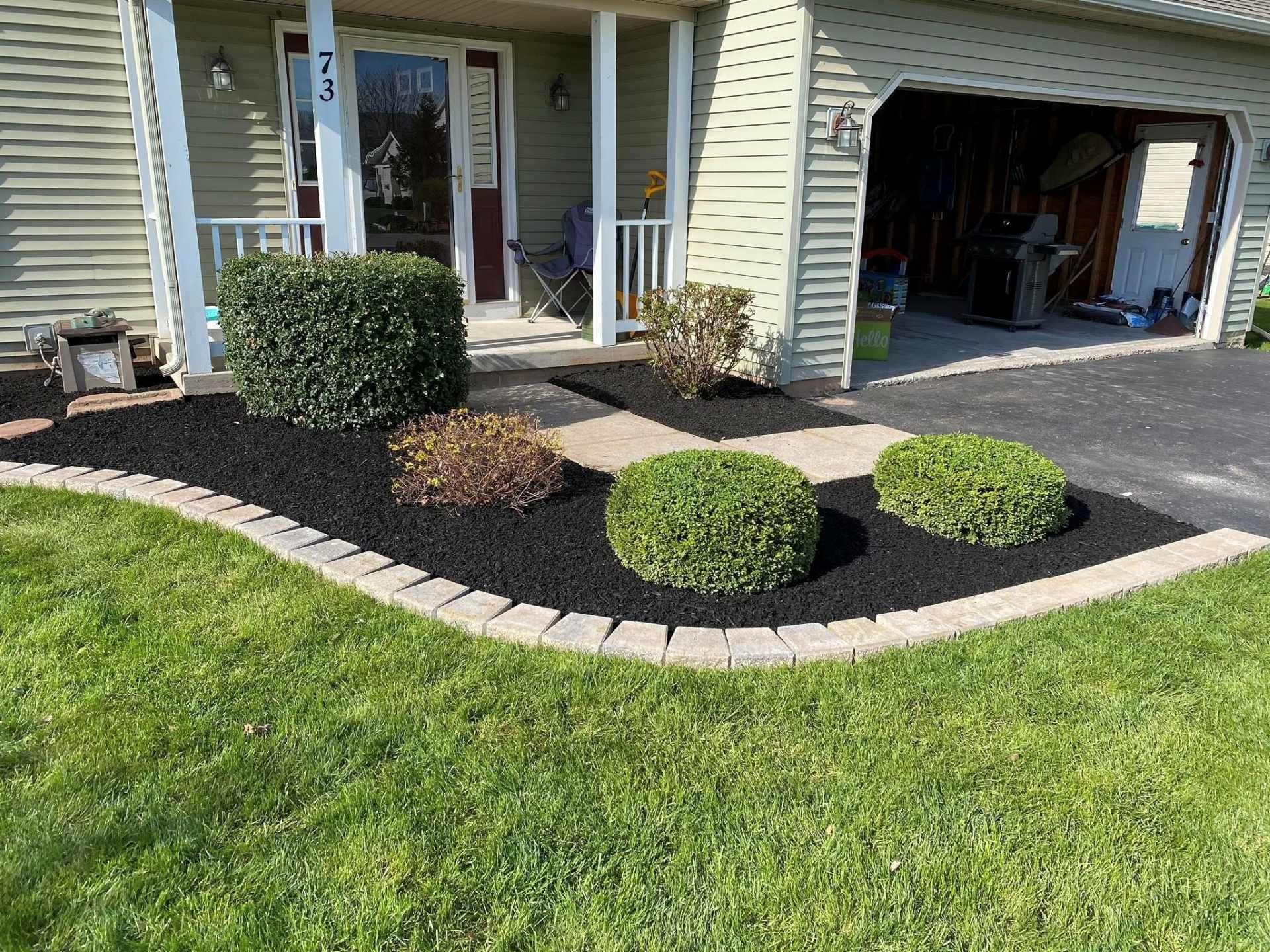 A front yard garden bed with black mulch, a stone border, and several manicured green shrubs in front of a house.