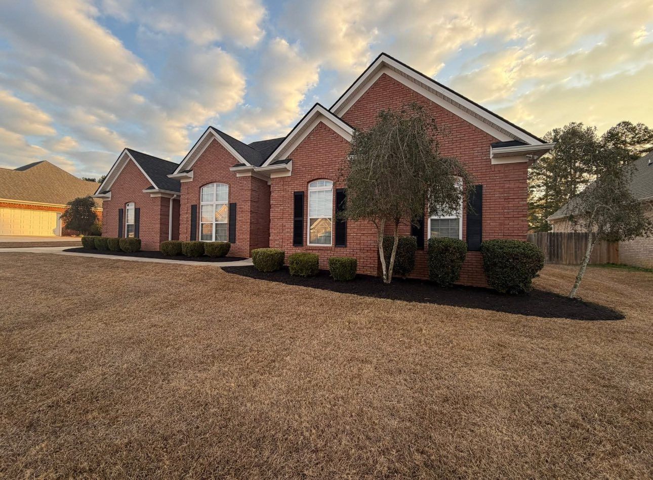 A one-story red brick house with a dark roof and manicured foundation shrubs under a cloudy sunset sky.