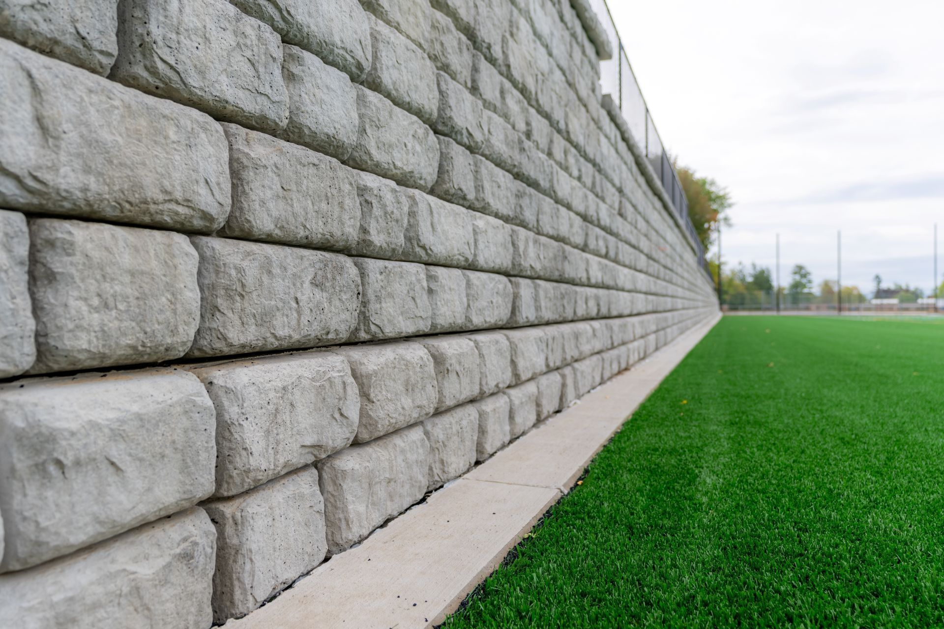 A low-angle view of a grey concrete block retaining wall beside a green synthetic turf field under an overcast sky.