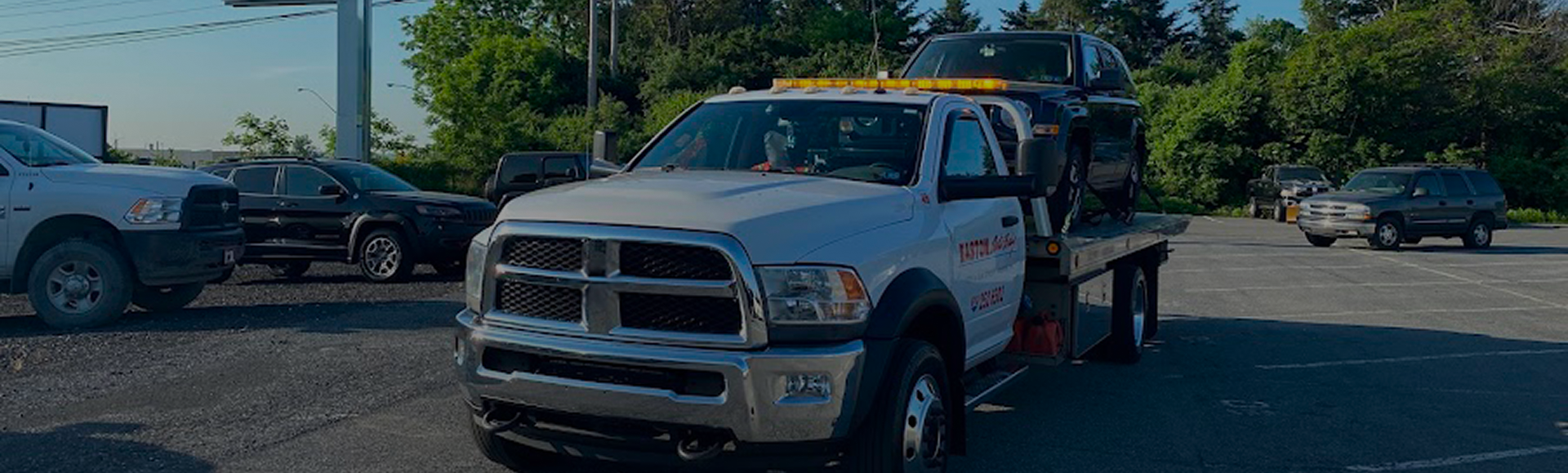White tow truck with emergency lights is in a parking lot, other cars and trees in the background. | Easton Auto Body