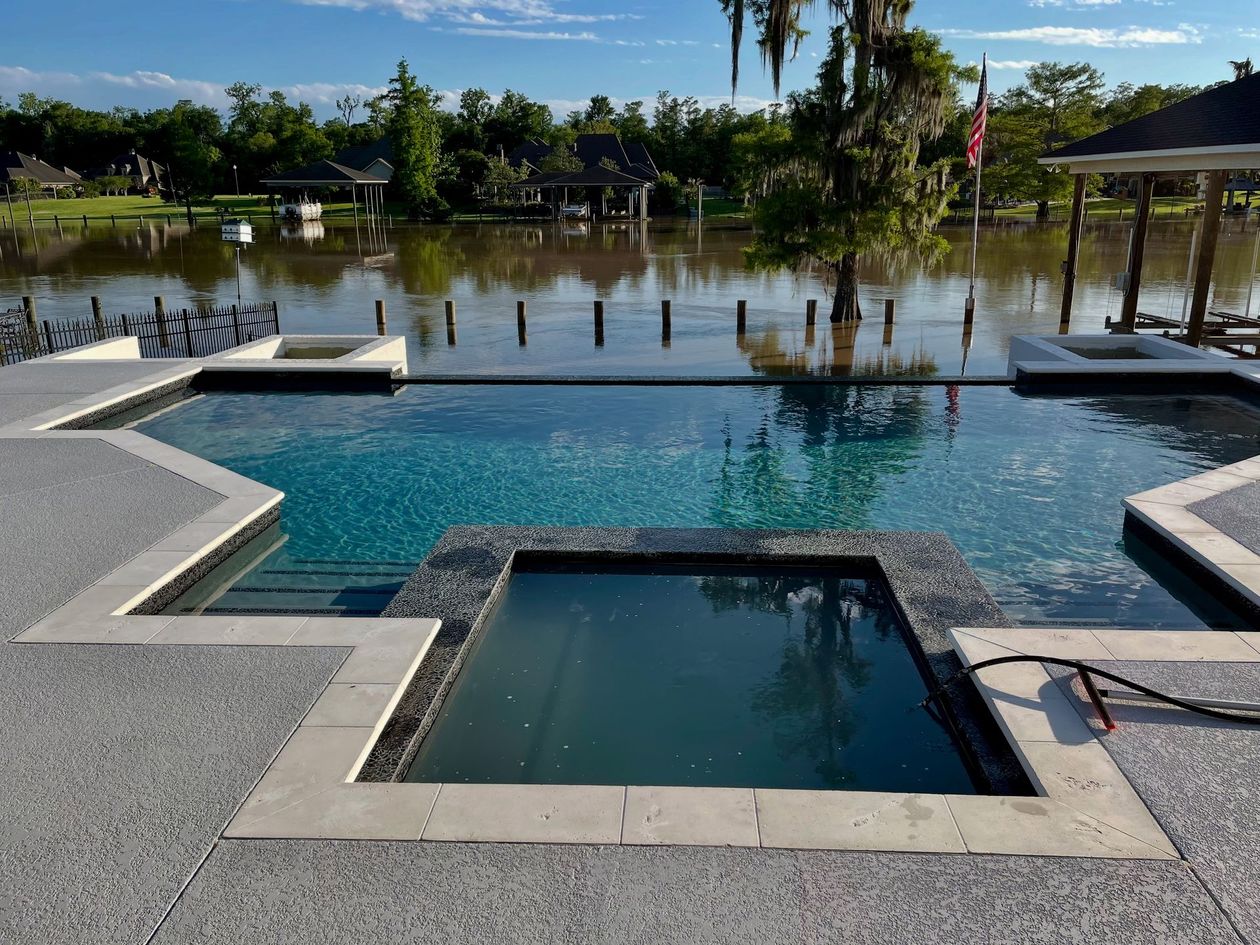 Pool with a dark-tiled hot tub and blue pool water, facing a lake under a blue sky, with houses in the background.
