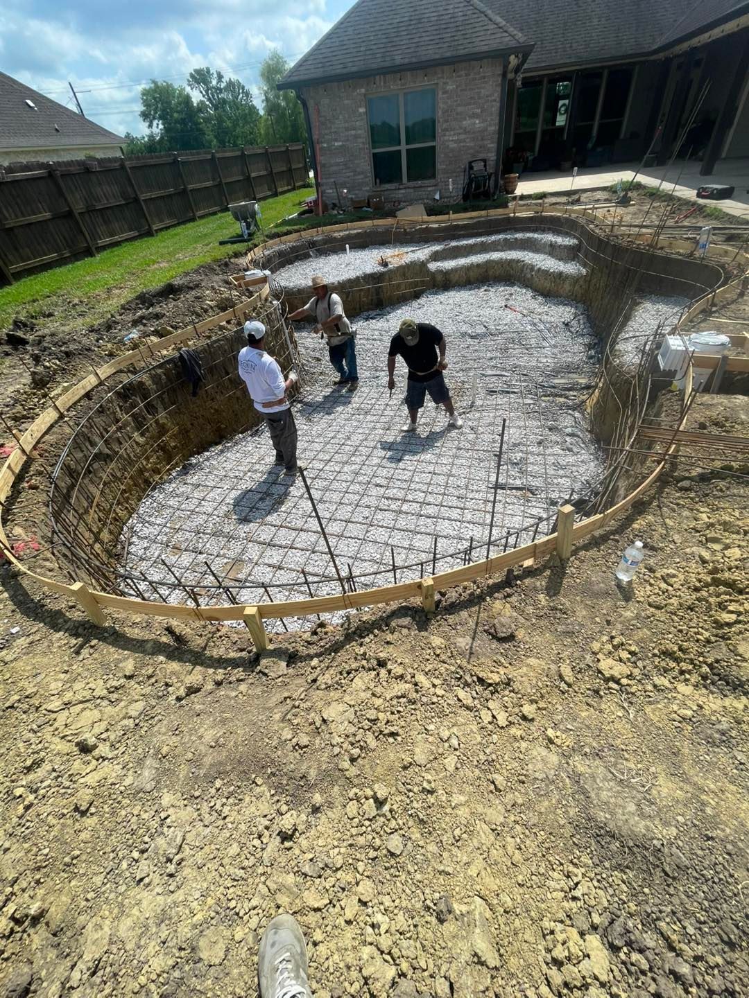 Construction of a pool: three workers inside an excavated pool shape, laying rebar on the pool's base. The setting is a backyard, with a house in the background.