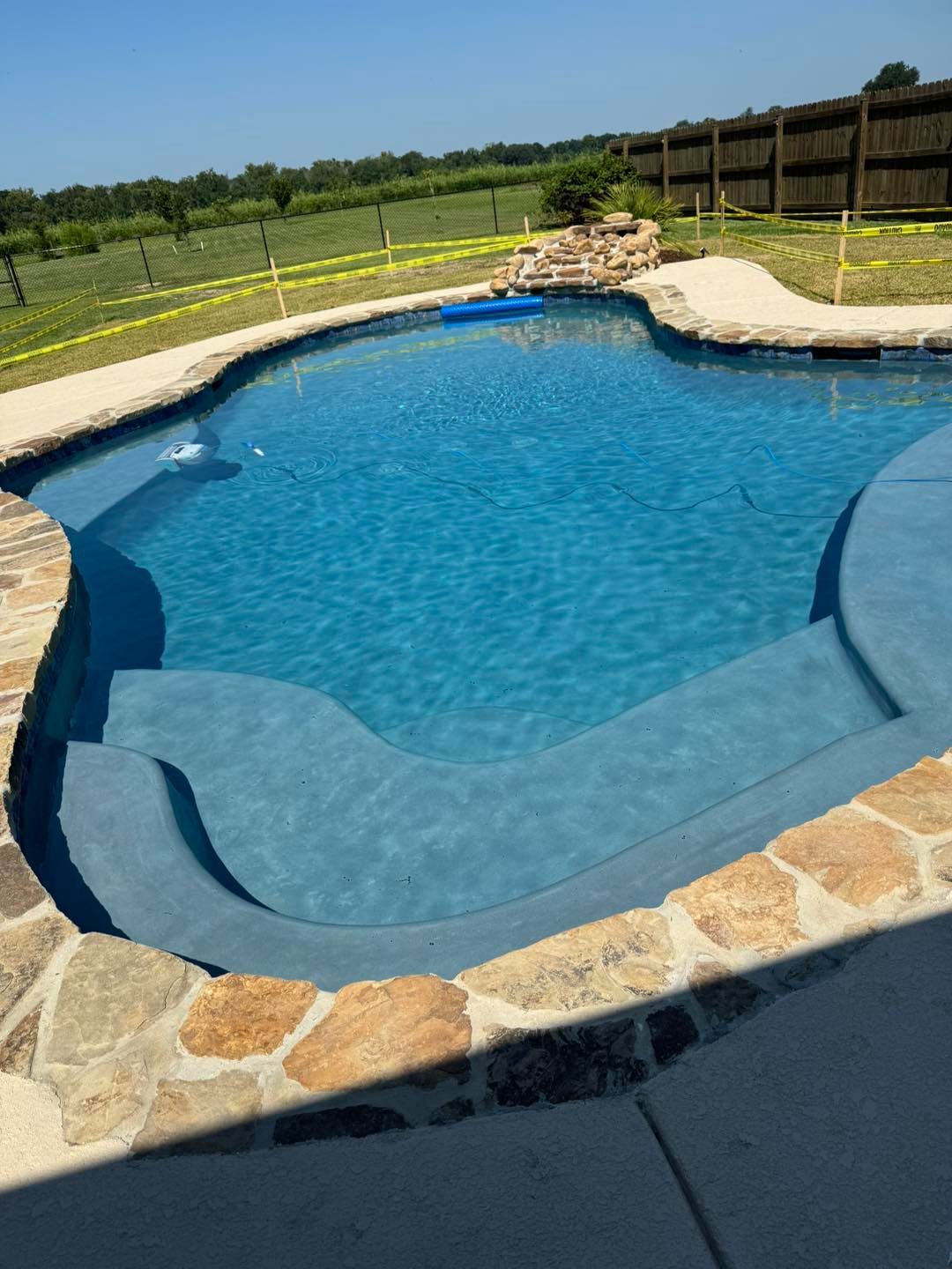 A blue-tiled swimming pool with stone edging, steps, and a rock waterfall feature. It is outdoors on a sunny day.