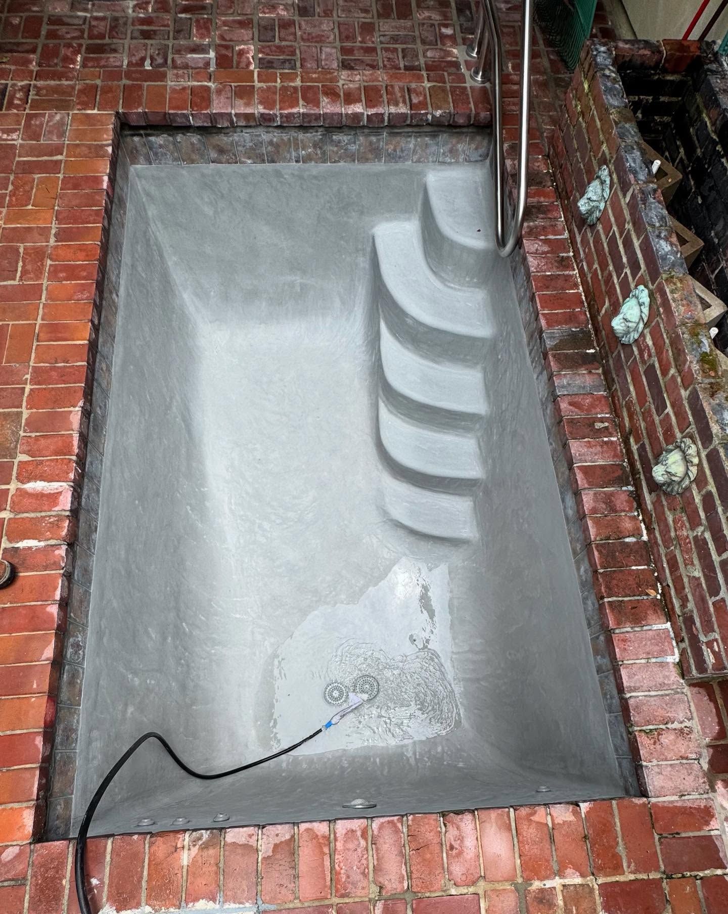Concrete pool being built, surrounded by red brick, with built-in steps and a metal handrail.