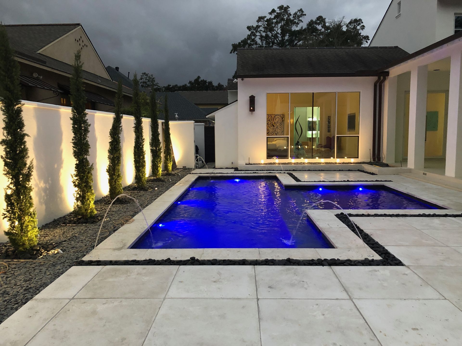 A modern backyard with a blue-lit pool, patio, and illuminated wall. Tall trees line the wall, and a house with glass doors is in the background.