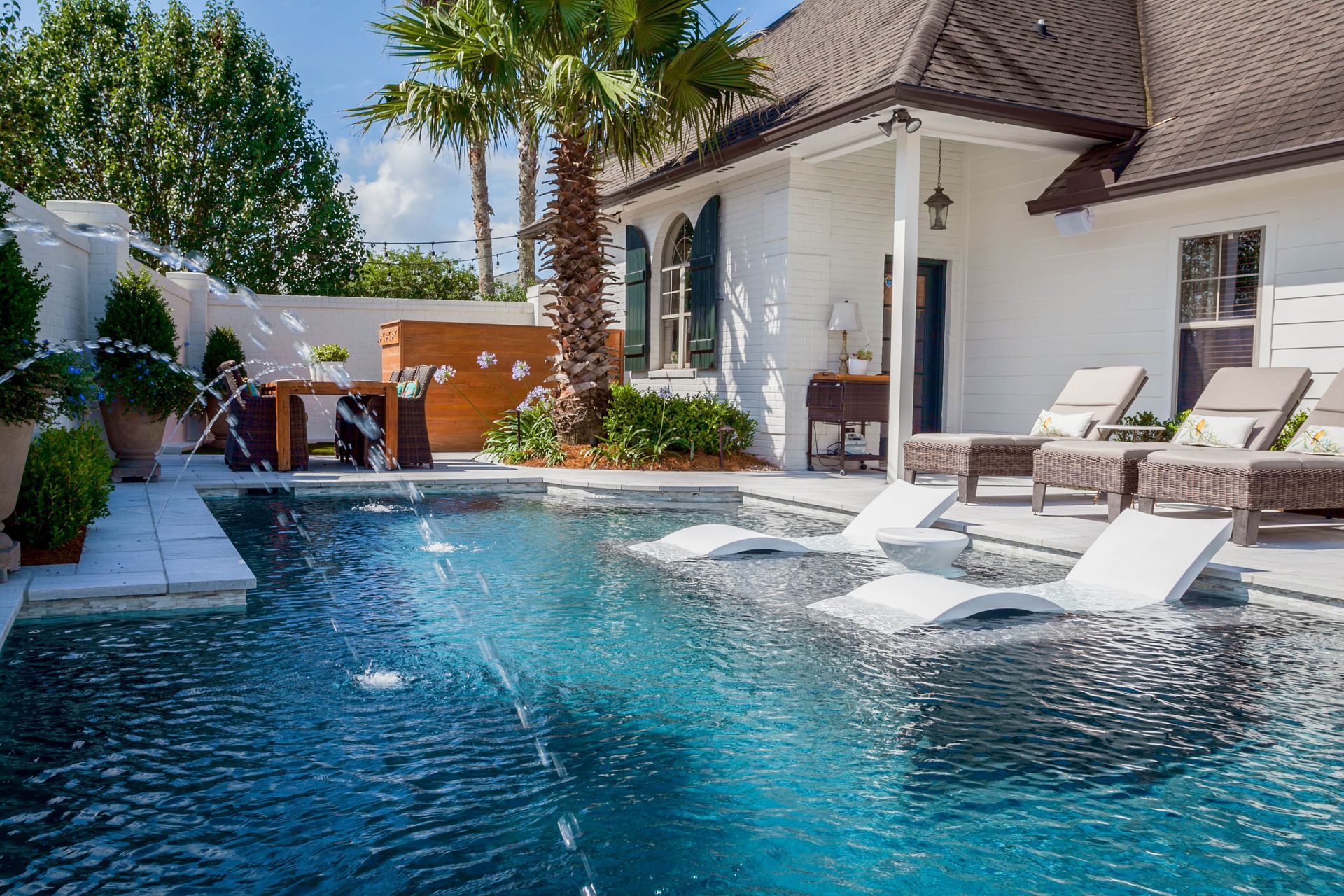Pool with white lounge chairs, palm trees, and a white house with dark shutters. Water fountains spray into the blue water.