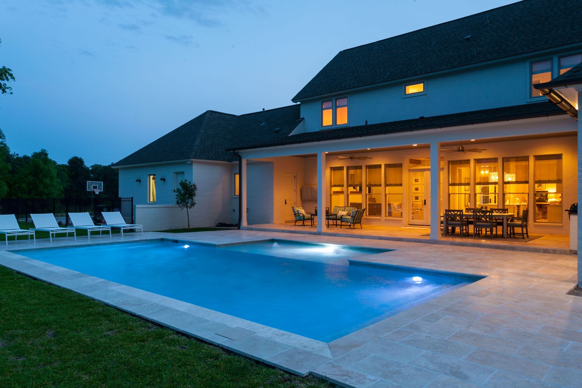 Backyard at dusk with pool and illuminated house. White lounge chairs by pool, outdoor dining and seating area on patio.