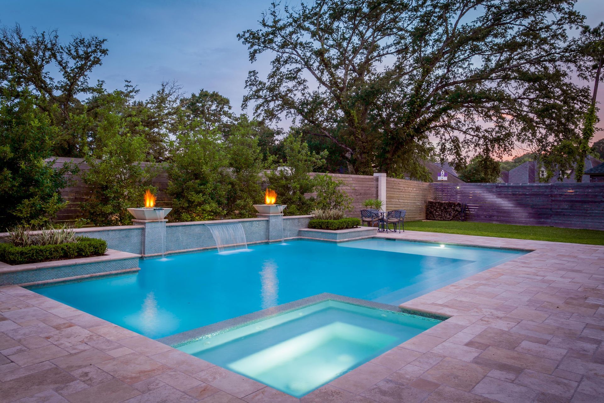 A luxurious backyard pool at dusk with a connected hot tub, waterfalls, and fire features. Lush greenery surrounds the area.