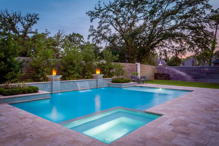 A luxurious backyard pool at dusk with a connected hot tub, waterfalls, and fire features. Lush greenery surrounds the area.