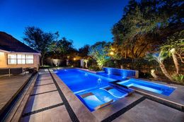 Nighttime view of a backyard pool with blue lighting and a spa. Concrete patio surrounds the pool, and trees are lit with spotlights.