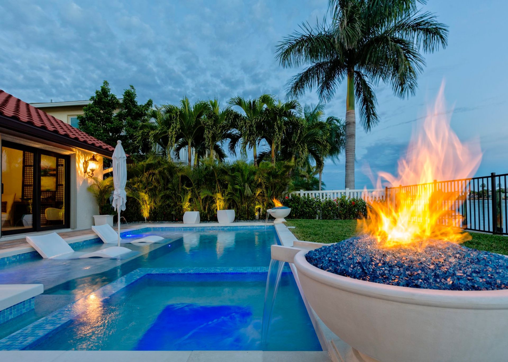 A luxurious outdoor pool area at dusk.  A fire pit glows with blue glass, palms and a house in the background.