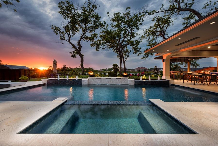 A small, illuminated backyard pool with a rock waterfall feature. The pool has blue water and the waterfall is made of brown rocks.