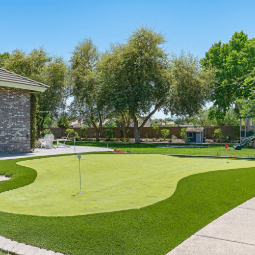 Backyard putting green with a flag, surrounded by lush green artificial turf. Trees and blue sky in the background.