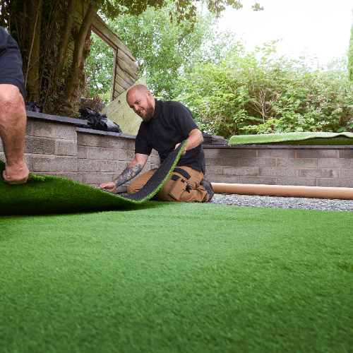Two men installing green artificial turf in an outdoor setting. One kneels, rolling out the turf near a brick wall, while the other holds the edge.