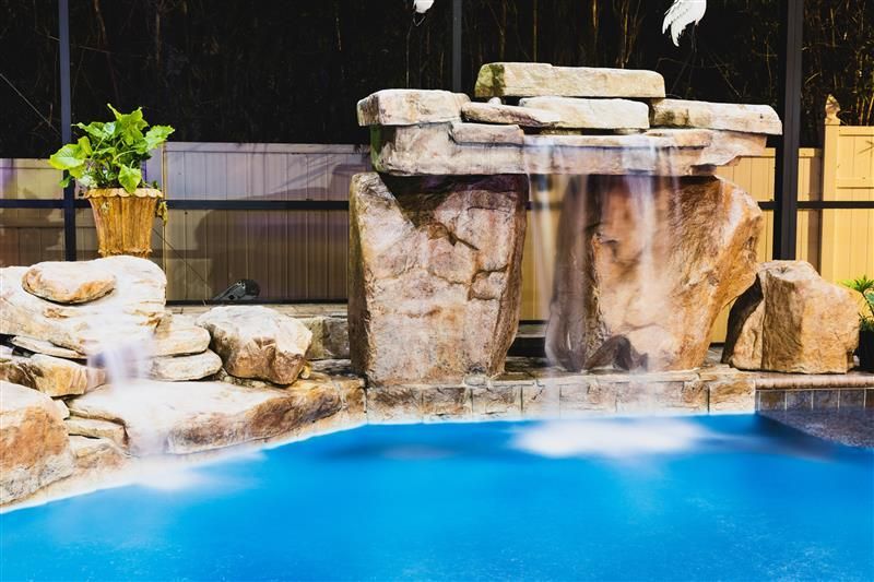 A small, illuminated backyard pool with a rock waterfall feature. The pool has blue water and the waterfall is made of brown rocks.
