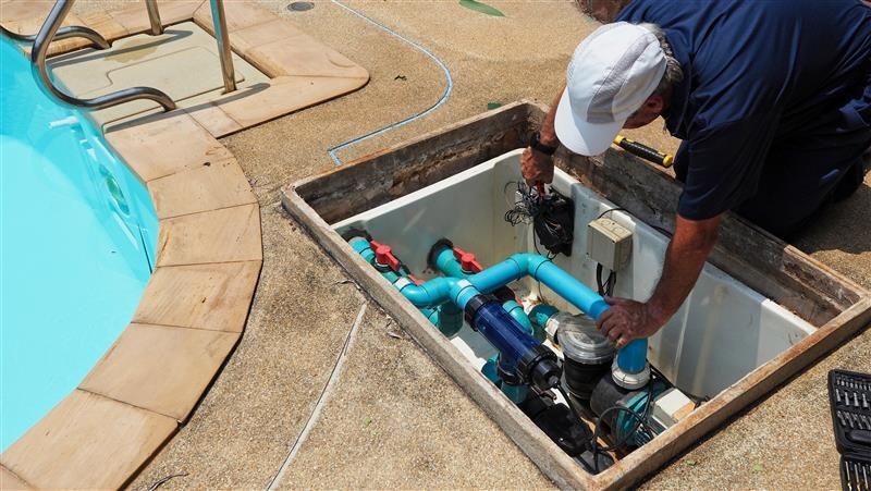 A man in a white cap works on pool equipment within an in-ground housing. The equipment includes blue pipes and valves next to the pool.