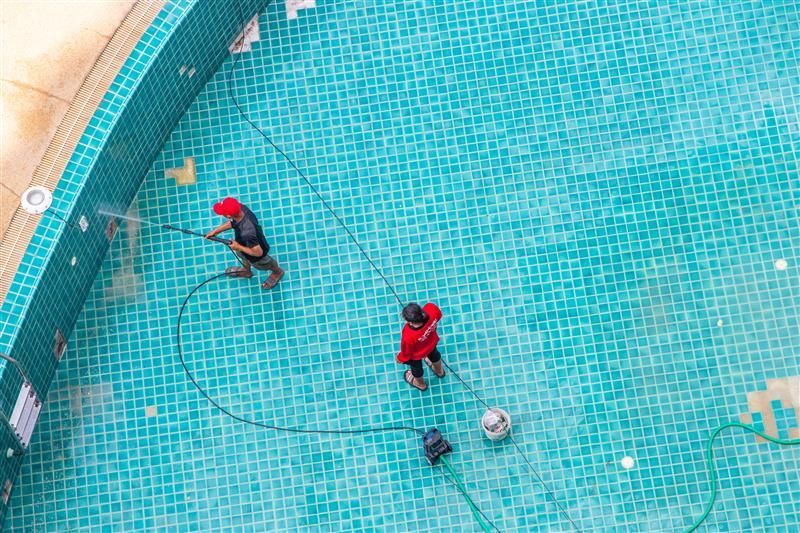 Two people in red shirts cleaning a turquoise-tiled swimming pool. They're using hoses and equipment near the pool's edge.
