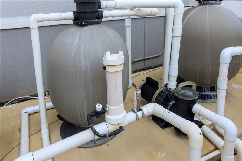 Pool filter and pump system with white PVC pipes, tan filters, and a black pump, outdoors.
