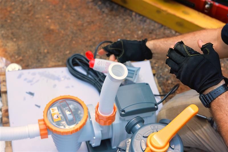 A person wearing black gloves assembles pool equipment with a pump and pipes, outdoors.