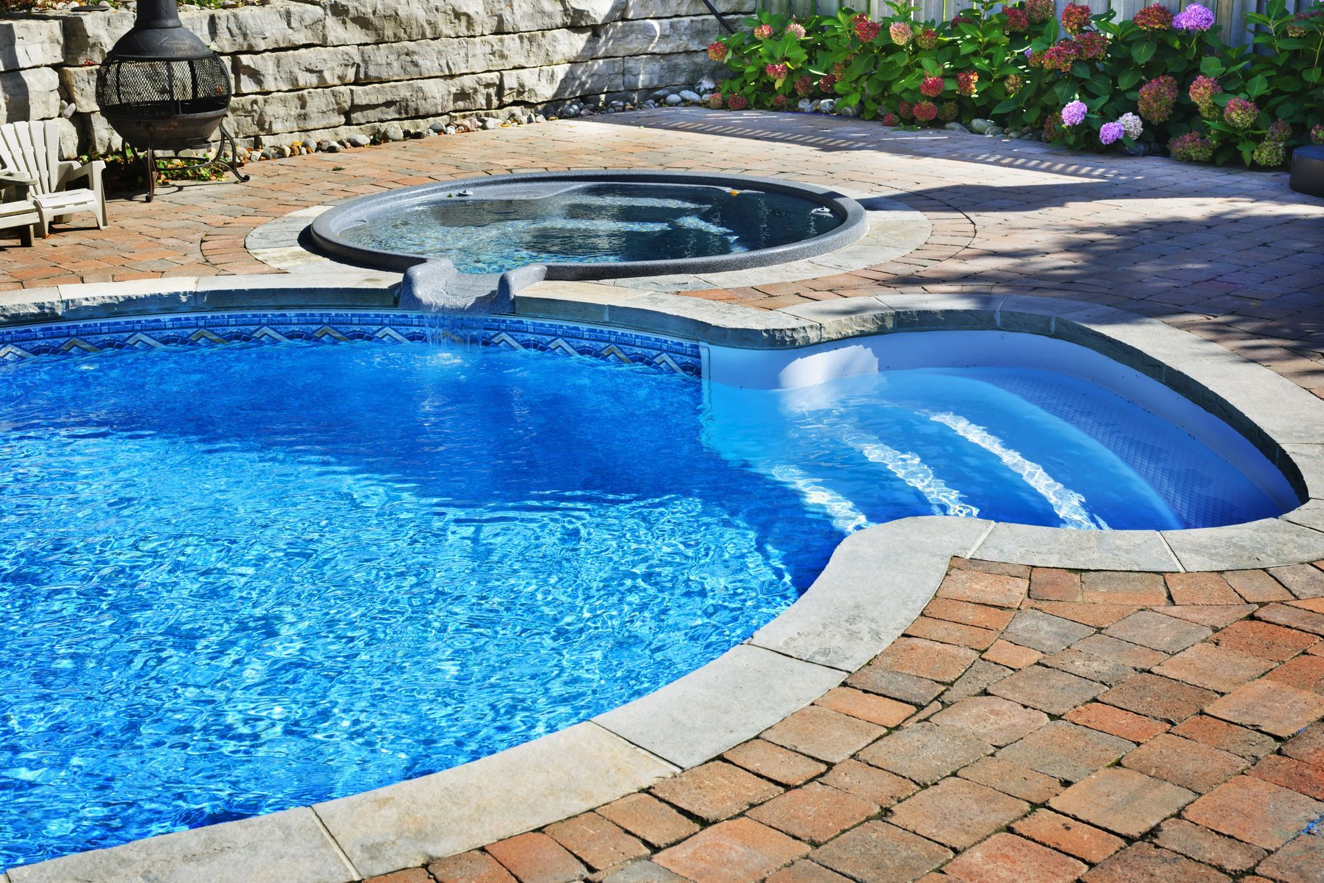 A backyard pool with a connected hot tub, surrounded by brick paving. The pool is filled with blue water and has steps.