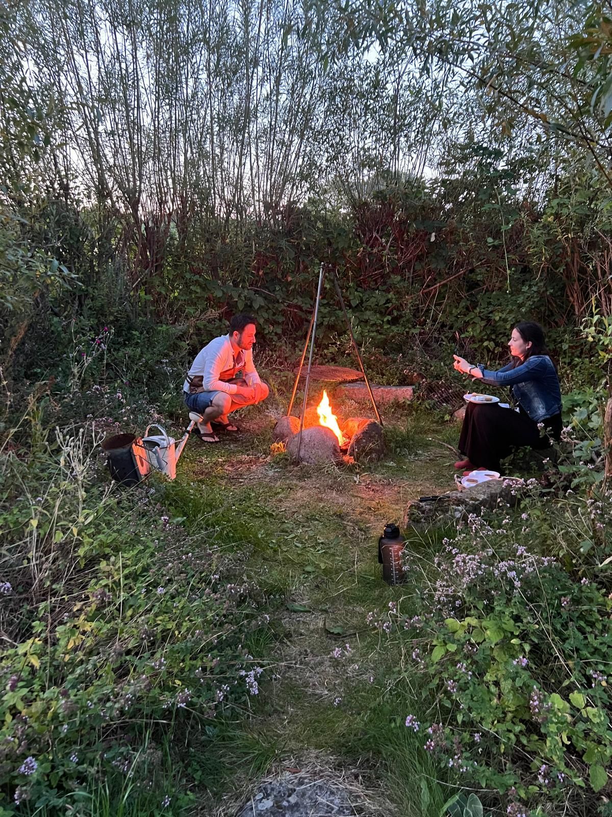 Two people are sitting around a campfire in the woods.