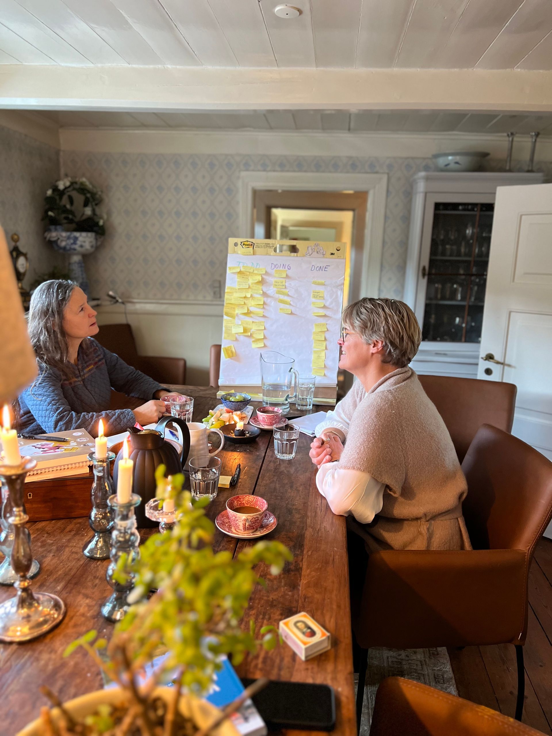 Two women are sitting at a table with a whiteboard on it.
