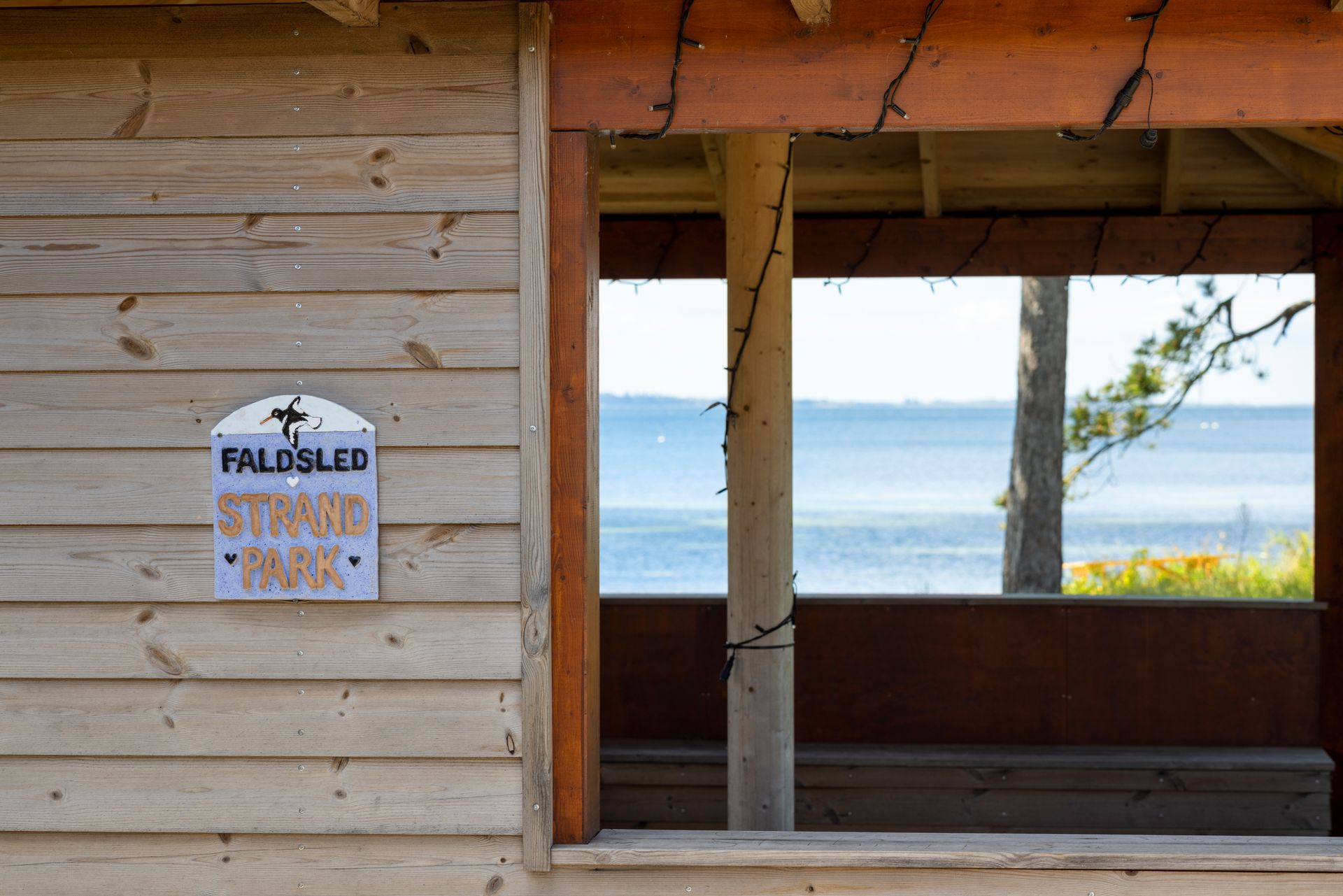 Cozy weathered beach hut façade with a ‘Falsled Strand Park’ sign, framed by a glimpse of the shoreline beyond