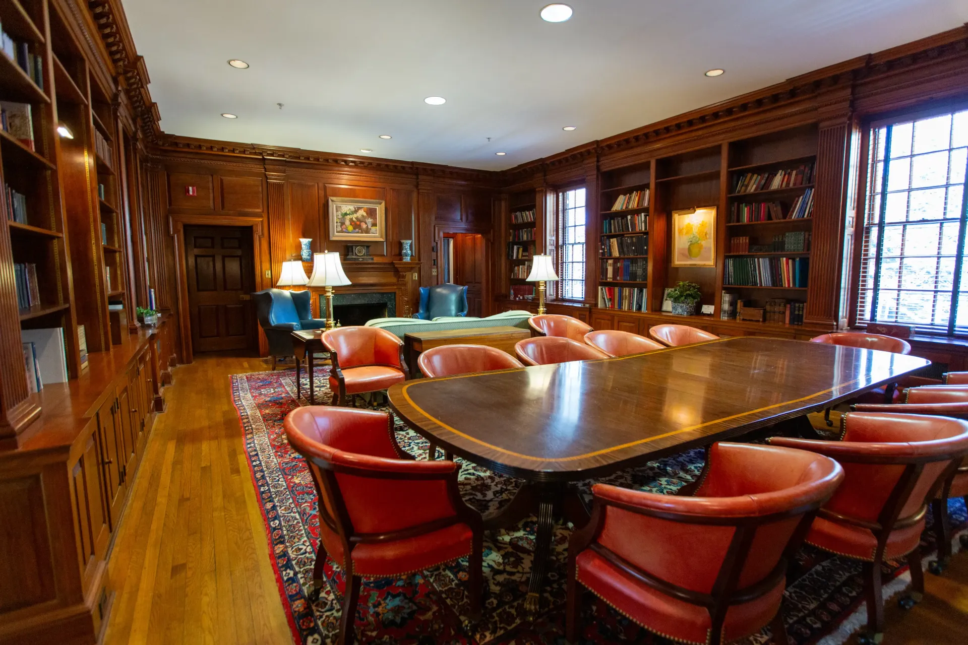 A wood-paneled library with a large conference table surrounded by red chairs, and bookshelves filled with books.