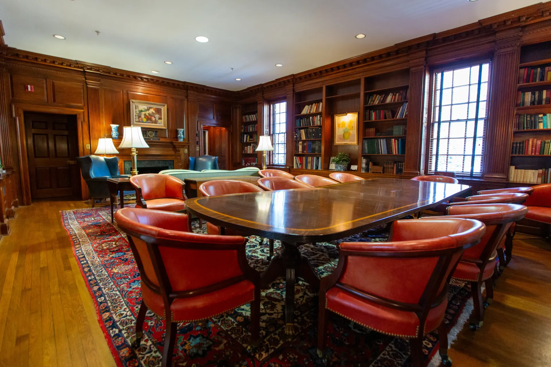 A wood-paneled library with a large table surrounded by red chairs, bookshelves, and a fireplace.