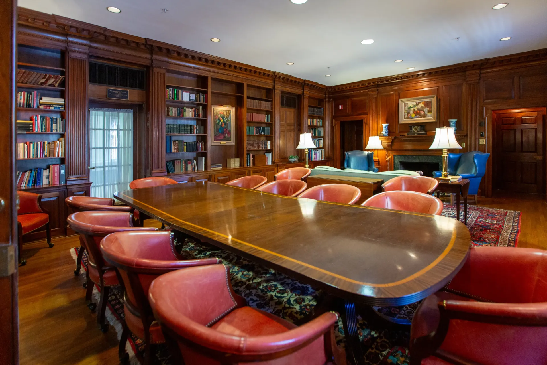 Library with a large table and red chairs surrounded by bookshelves and wood paneling.