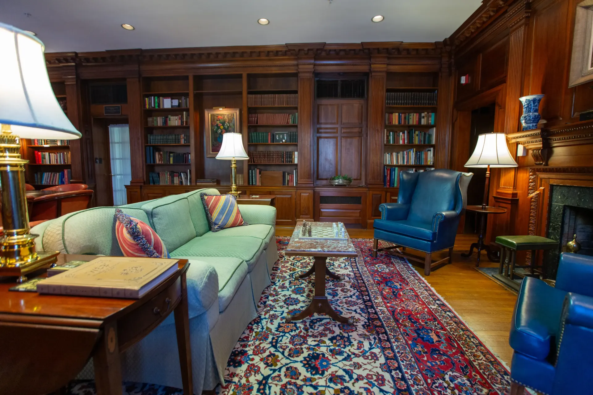 Cozy library with wood paneling, green couch, blue chairs, oriental rug, and bookshelves filled with books.