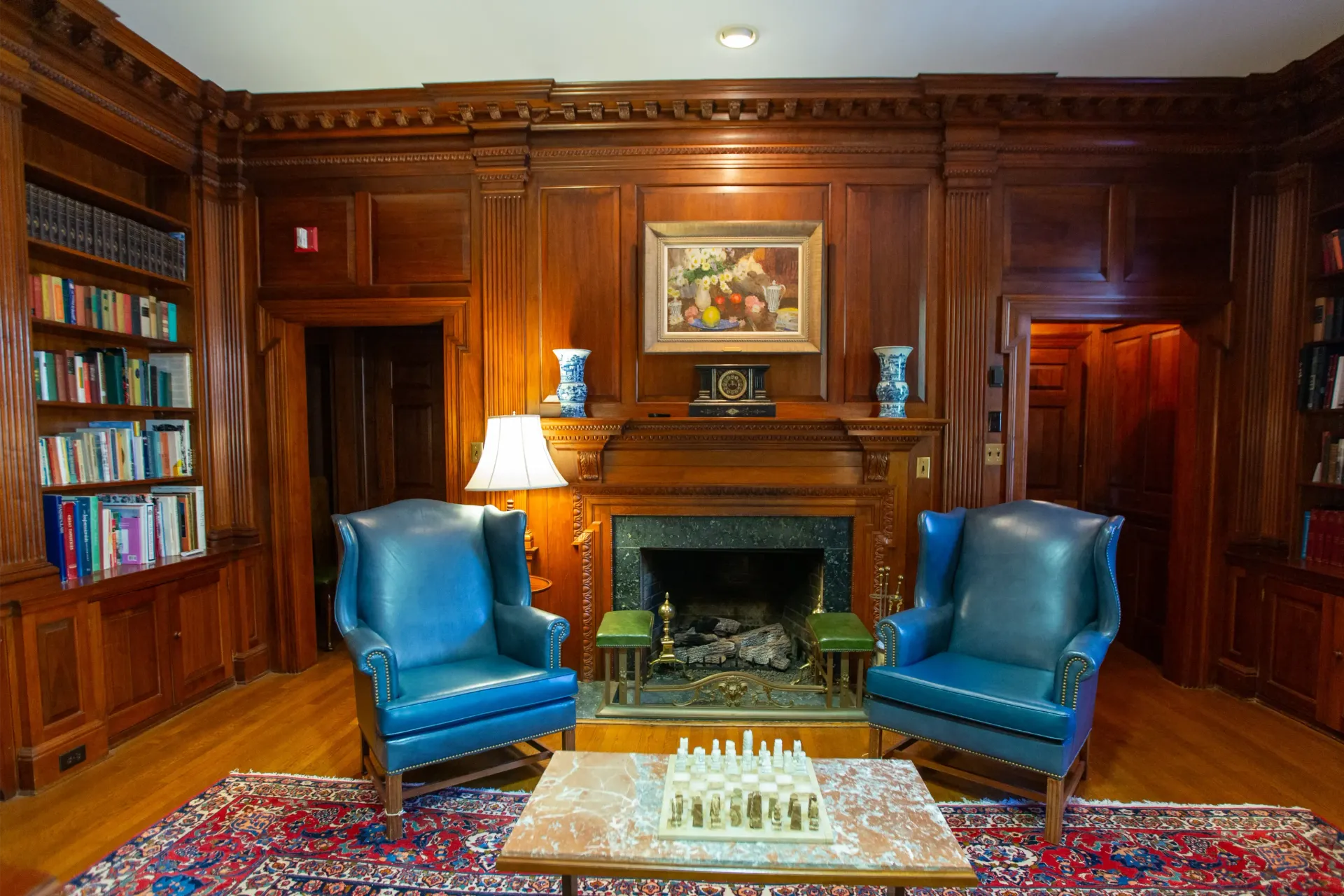 Cozy library with wood paneling, two blue chairs facing a fireplace, and bookshelves.