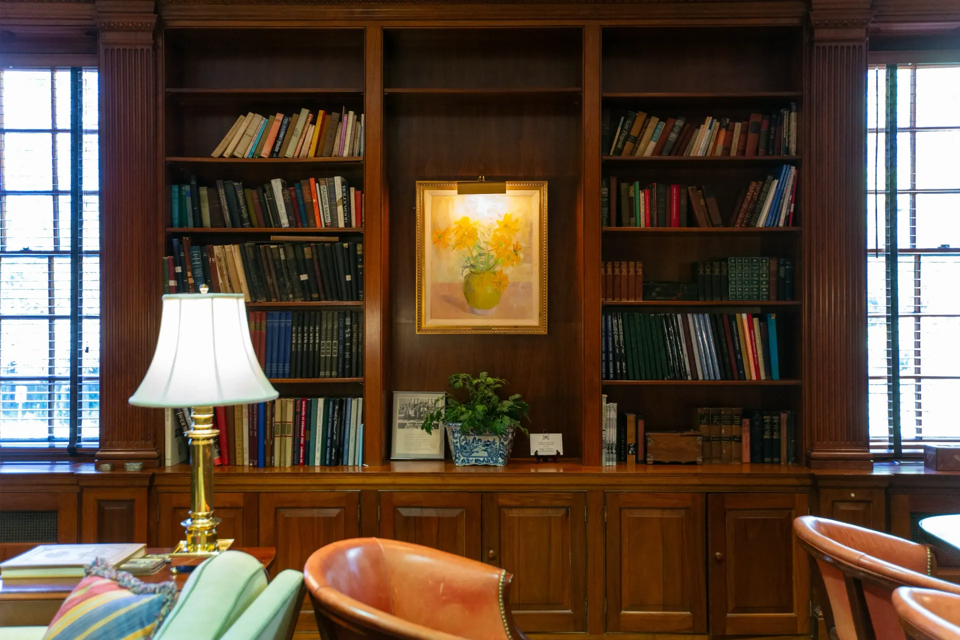 Wooden bookcase filled with books, a painting, and a lamp. Two armchairs are in the foreground.