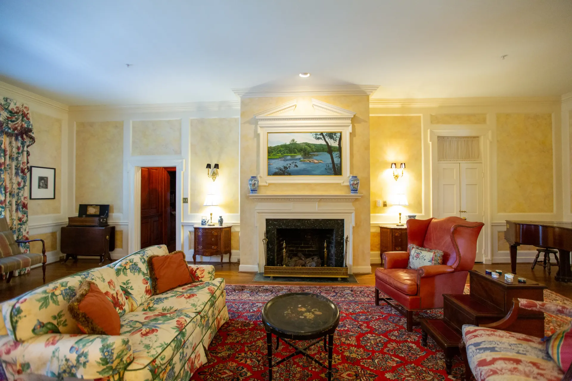 Cozy living room with floral sofa, red rug, fireplace, and framed landscape artwork on the wall.