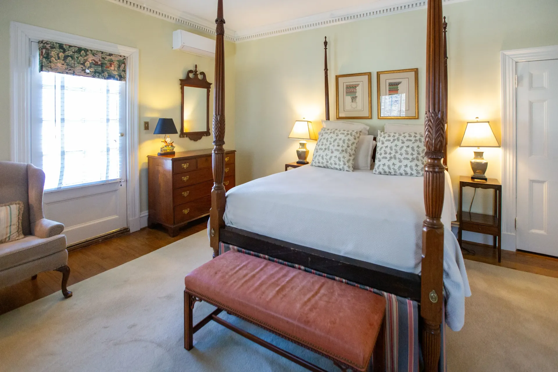 Bedroom with four-poster bed, bench, dresser, armchair, and lamps; light green walls, pale rug, and window with floral shade.