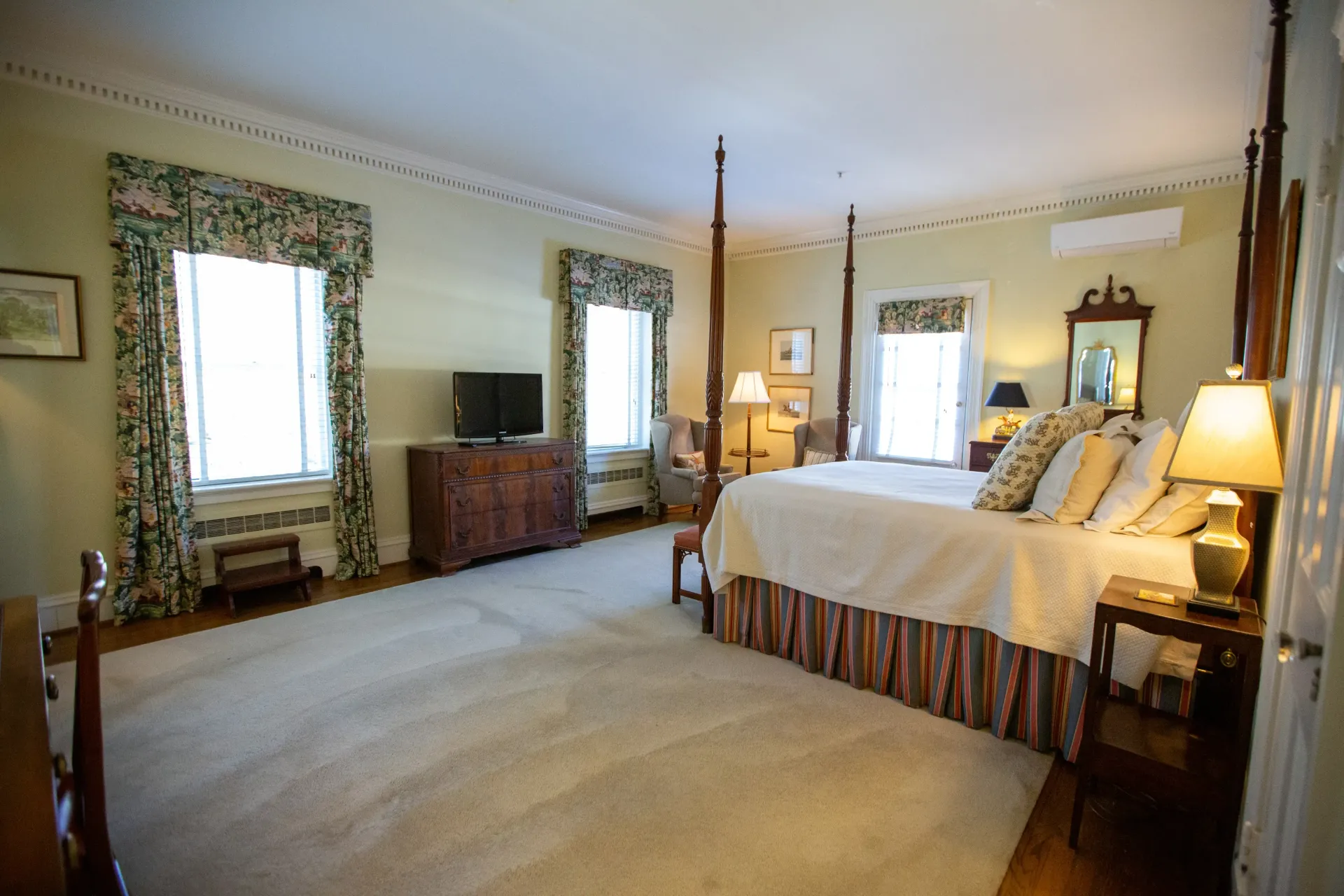 Spacious bedroom with a four-poster bed, rug, windows with floral curtains, and a TV.