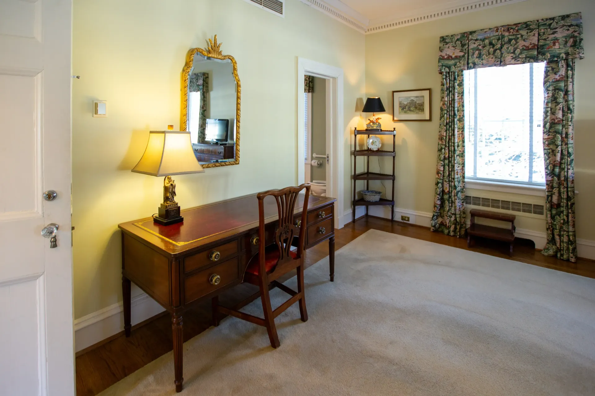 A bedroom with a desk, mirror, window, and small bookshelf, green walls, and light carpet.