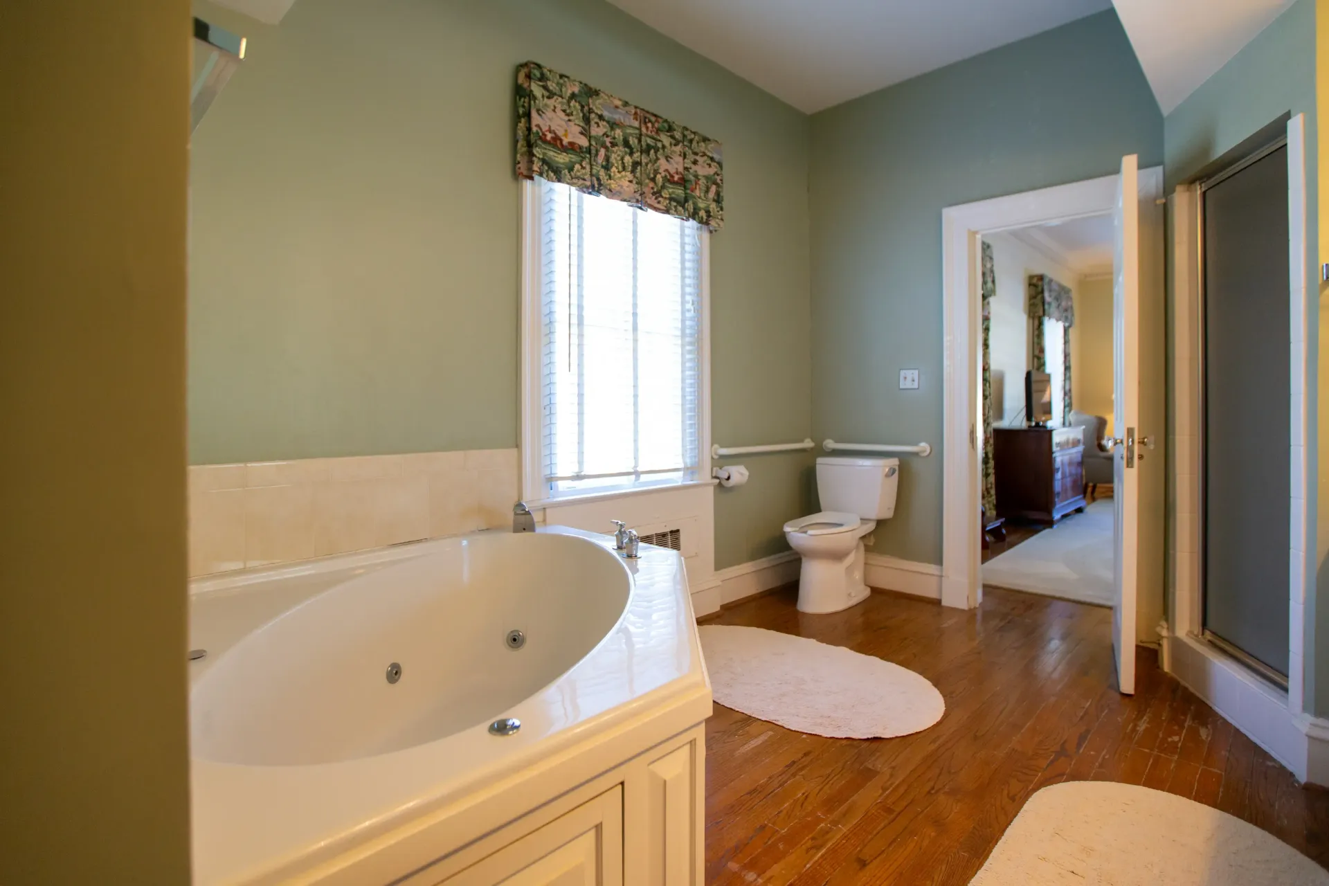 Bathroom with a white jacuzzi tub, toilet, and shower. Green walls, wood floor, open doorway.