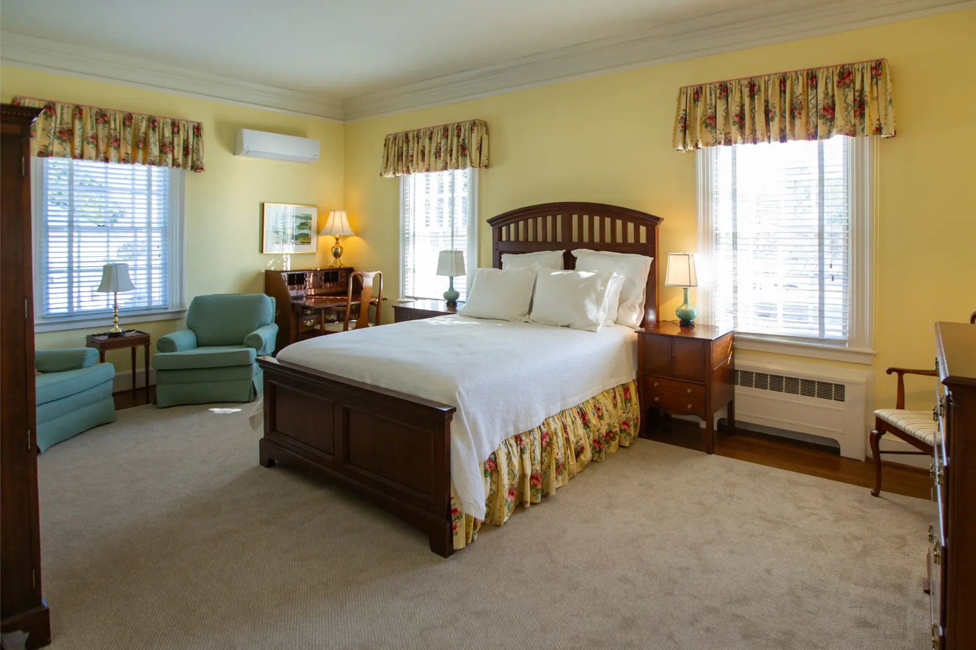 Bedroom with queen bed, two armchairs, desk, and two windows. Yellow walls and beige carpet.