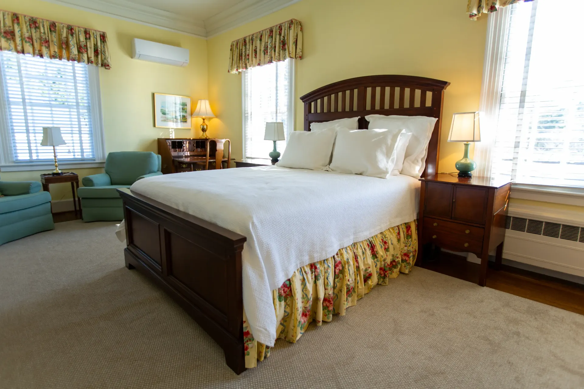 Bedroom with king bed, floral bed skirt, windows, two chairs, and yellow walls.
