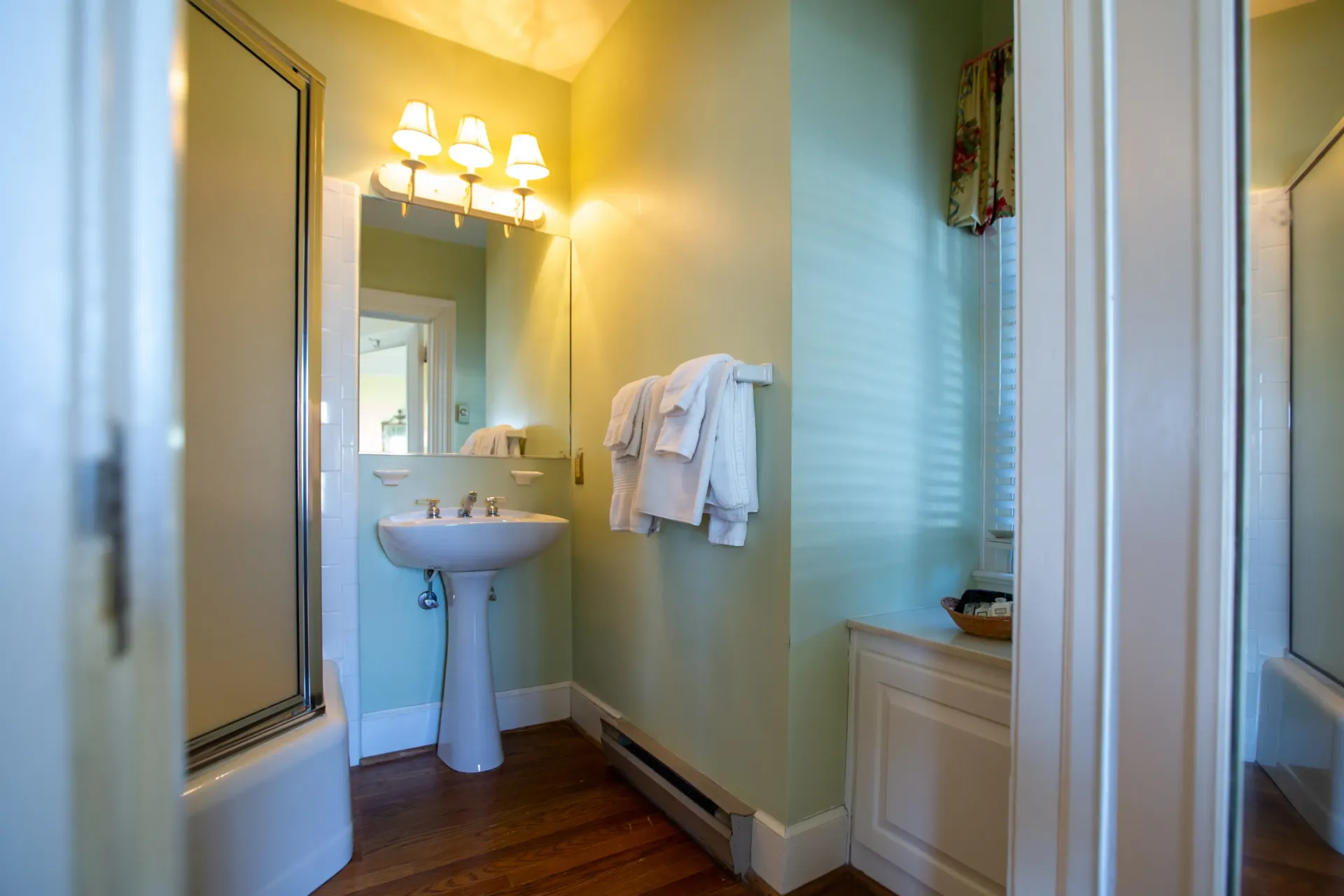 Small bathroom with a pedestal sink, mirror, shower, and white towels on a rack. Soft green walls, wooden floor.