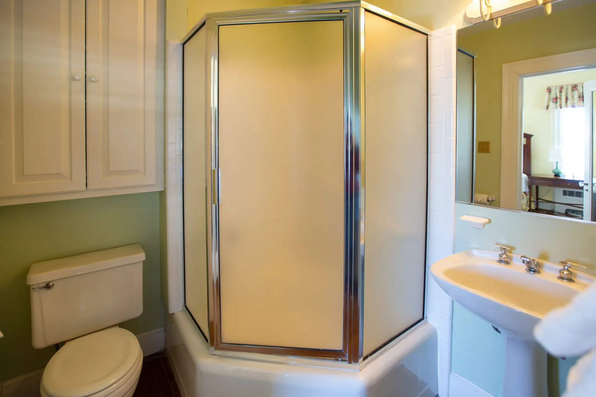 Bathroom with a corner shower, toilet, sink, and cabinet. Green walls, white fixtures, and a mirror are visible.