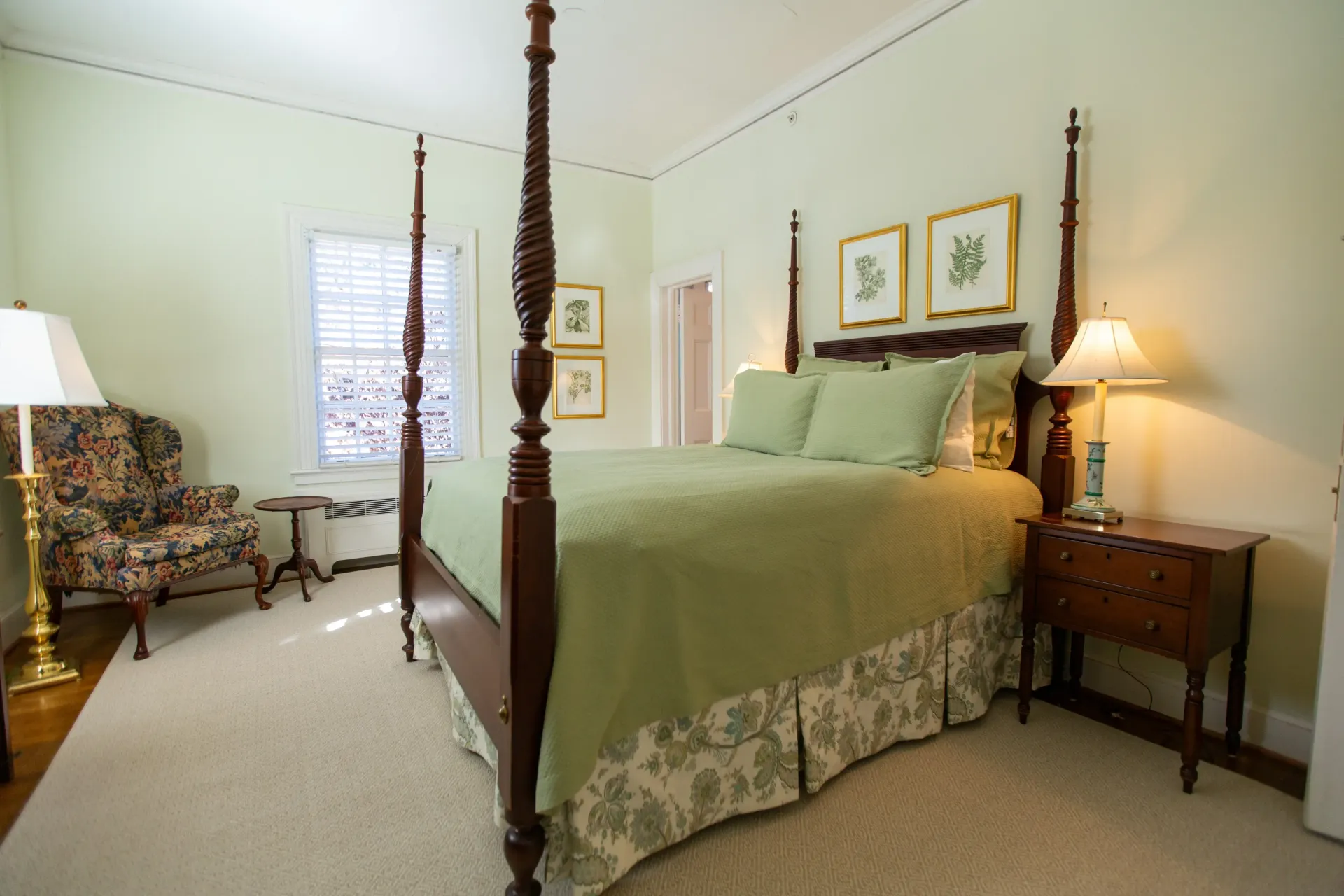 Bedroom with four-poster bed, green bedding, floral patterned chair, nightstand, and framed botanical prints.