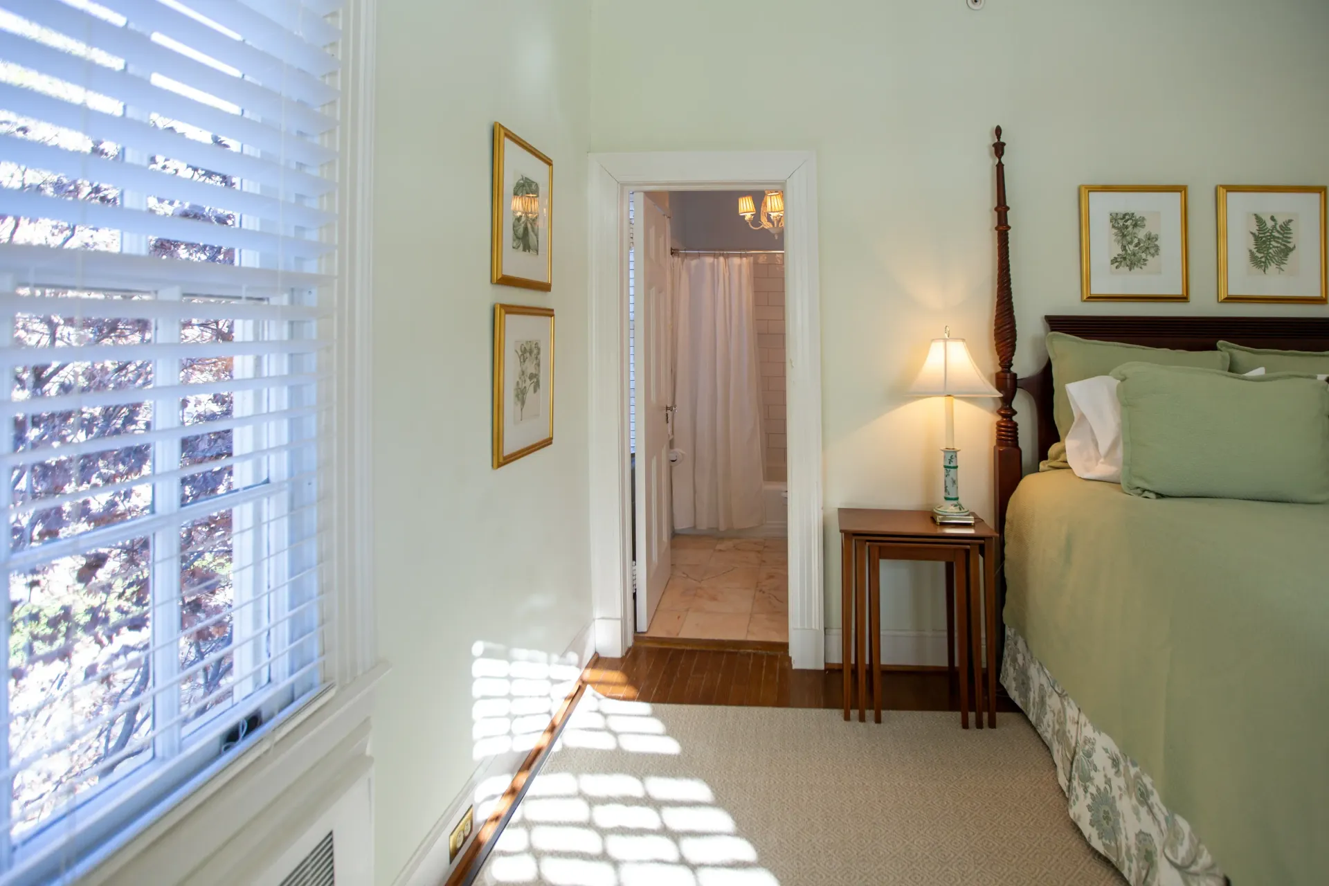 Bedroom with pale green walls, four-poster bed, window with blinds, and doorway to a bathroom.
