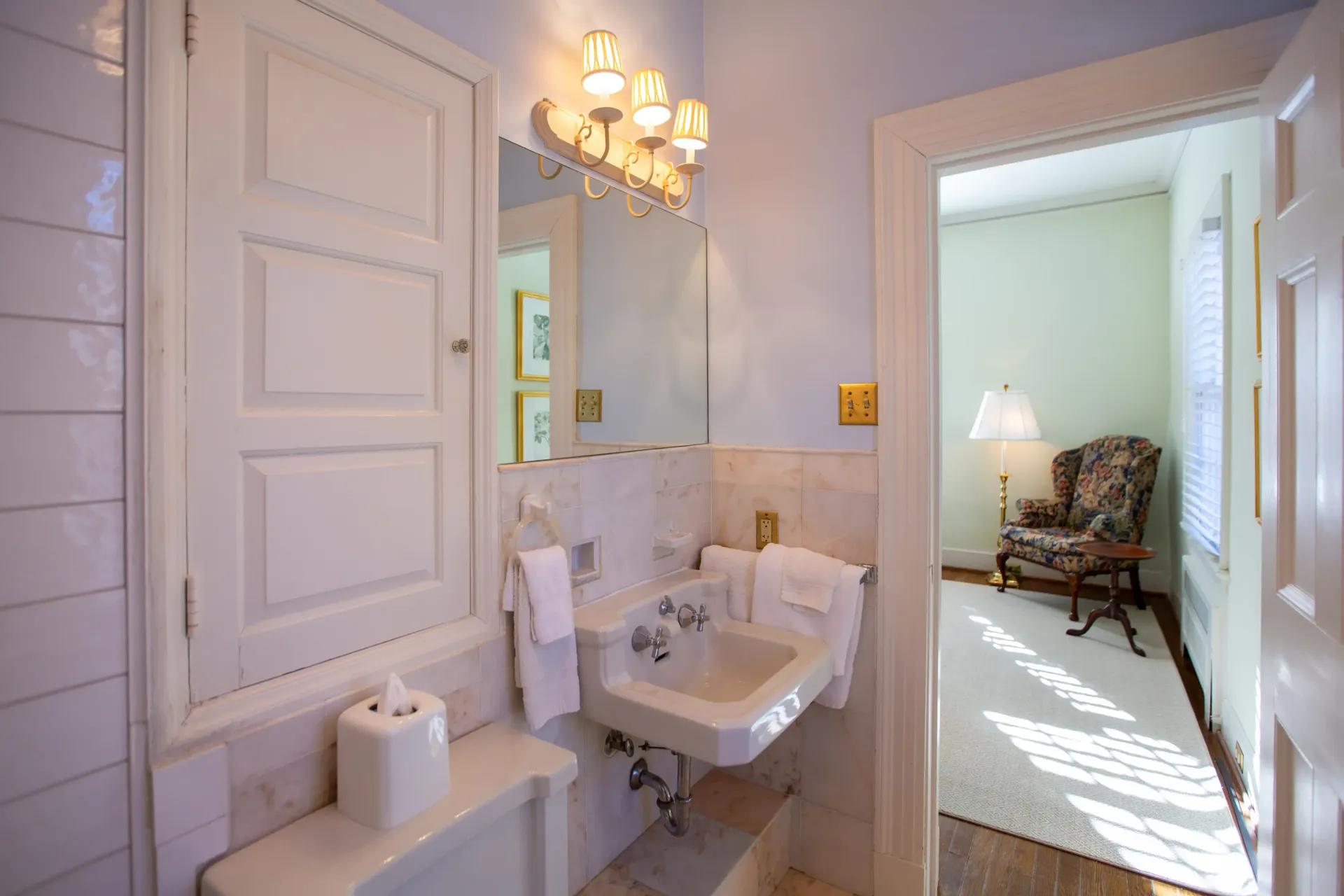 Bathroom with a white sink, mirror, and a doorway leading to a room with a chair.