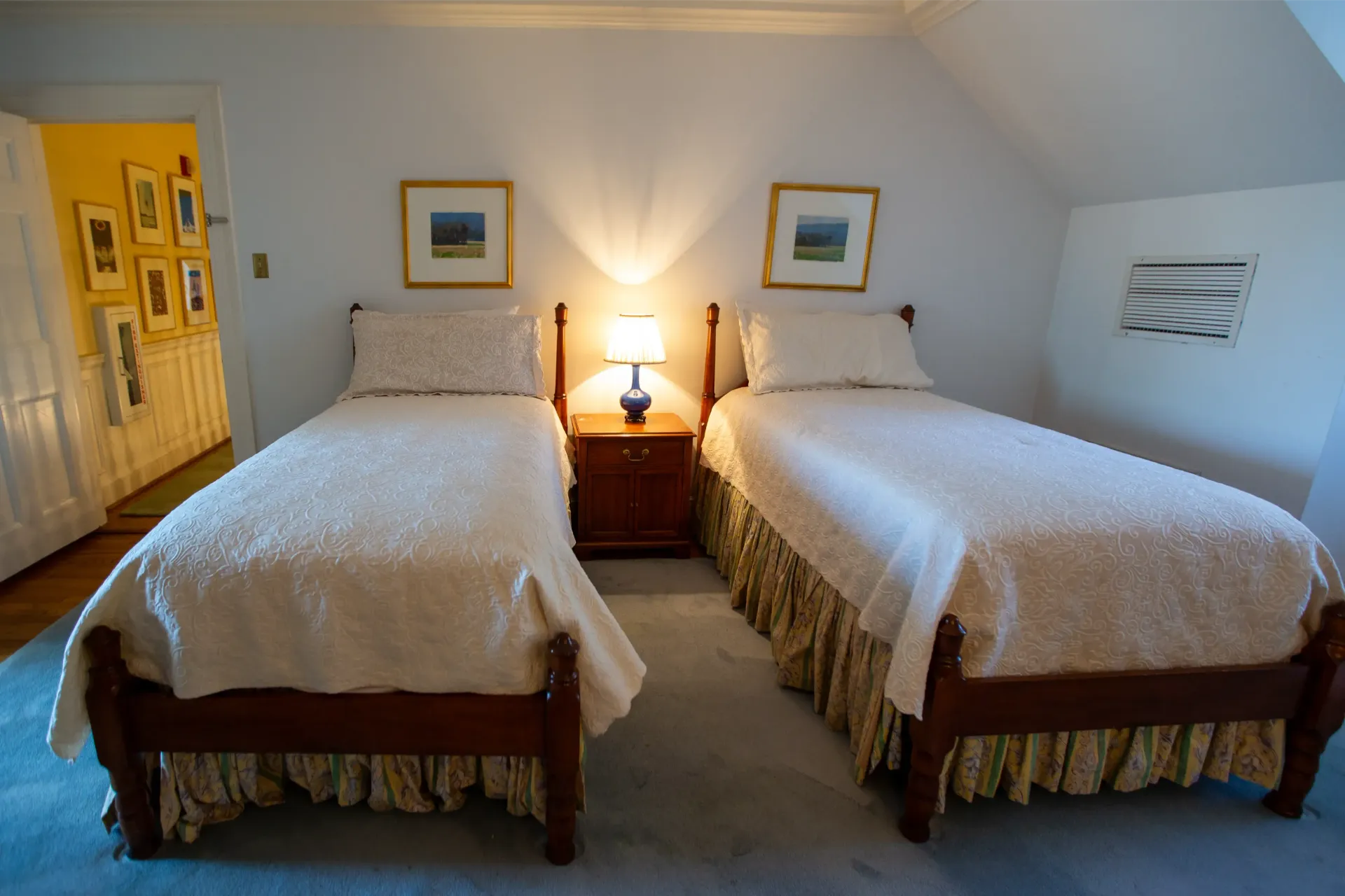 Two twin beds in a bedroom, with a nightstand and lamp between them. Wall art and a doorway are visible.